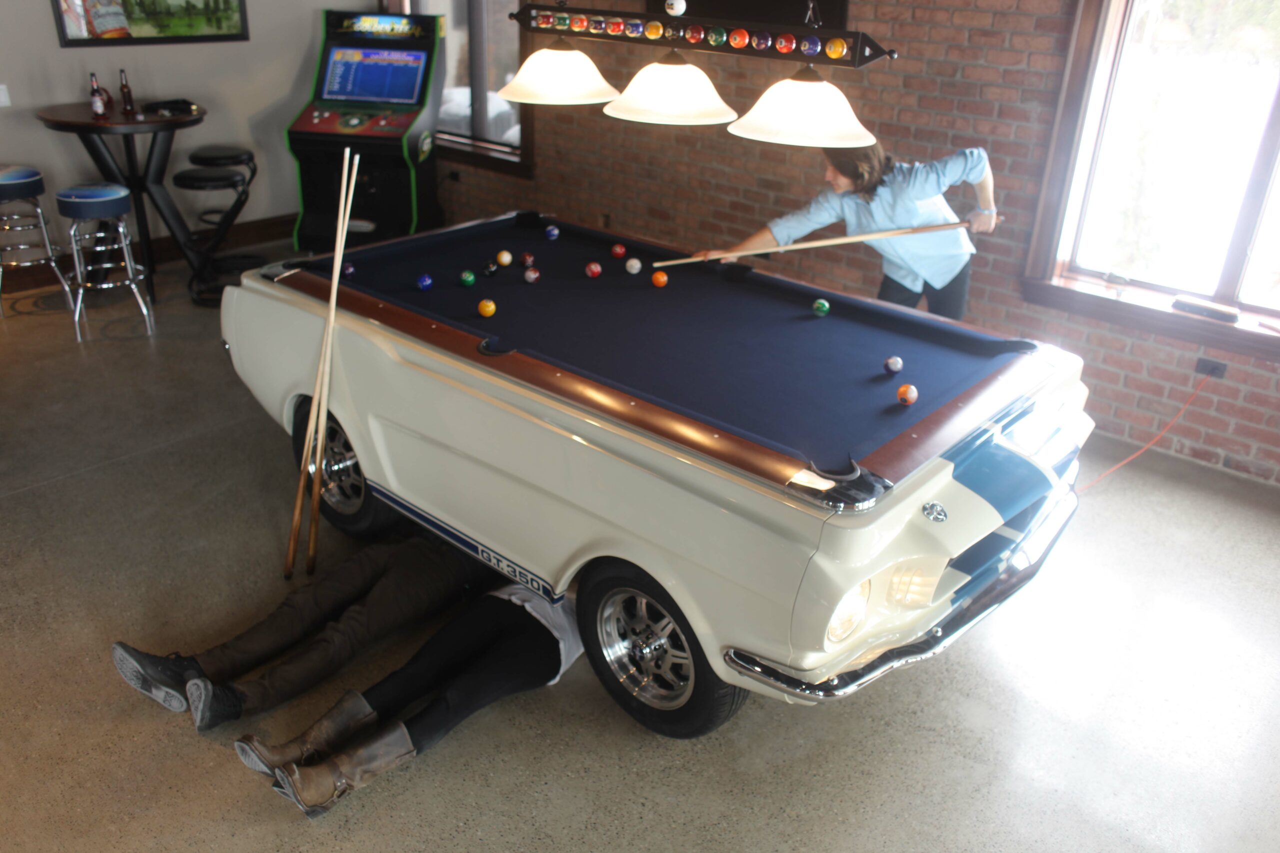 people pretending to be mechanics working under a Ford Shelby car pool table while another is taking a shot!